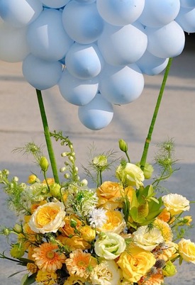 Basket of yellow white flowers with blue balloons cluster on top with leaves